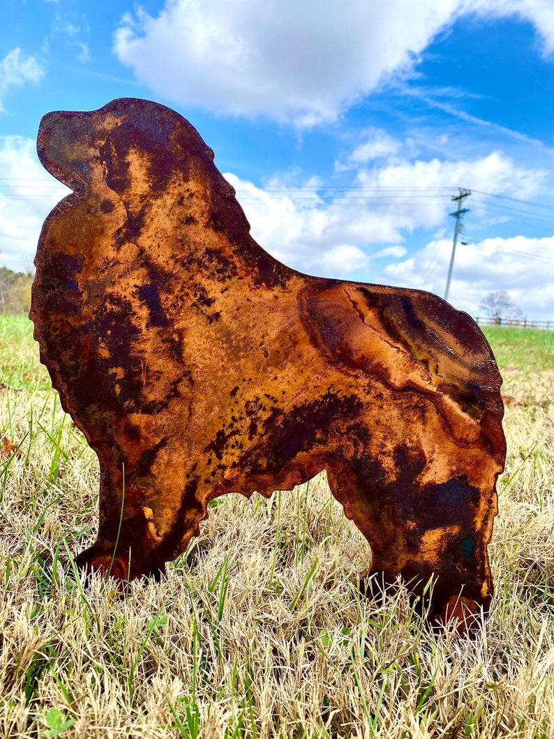 K&ouml;nnte beinhalten: Eine rostige Metallsilhouette eines Hundes, der auf einem Bett aus trockenem Gras steht. Der Hund ist nach rechts gerichtet, mit dem Kopf nach oben geneigt. Die Silhouette ist eine einfache Umrandung der Hundeform, ohne Details.