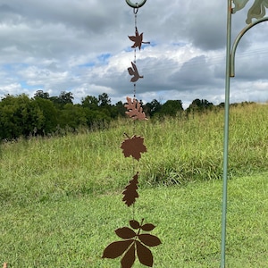 May include: A metal wind chime with a series of brown leaf cutouts hanging from a green metal hook. The leaves are of different shapes and sizes, and the chime is hanging in a grassy field.
