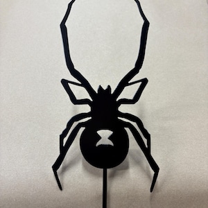 May include: A black metal spider silhouette topper featuring a stylized hourglass shape cut out from the abdomen. The spider has eight angular legs and is attached to a thin vertical metal stake extending from the bottom of the body.