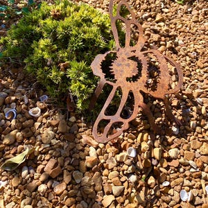 May include: A rusty metal butterfly garden sculpture with intricate cut-out details stands amidst small brown and tan pebbles. A vibrant green bush is to the left of the sculpture. The sculpture has a weathered appearance.
