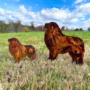 K&ouml;nnte beinhalten: Zwei rostige Metall-Hunds силуэты, eine gro&szlig;e und eine kleine, stehen auf einem grasbewachsenen Feld vor blauem Himmel mit wei&szlig;en Wolken. Die gr&ouml;&szlig;ere Silhouette ist von einem stehenden Hund mit dem Kopf zur Seite gedreht, w&auml;hrend die kleinere Silhouette von einem sitzenden Hund mit dem Kopf nach unten ist.