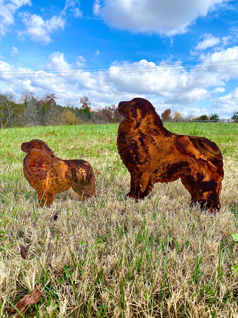 K&ouml;nnte beinhalten: Zwei rostige Metall-Hunds силуэты, eine gro&szlig;e und eine kleine, stehen auf einem grasbewachsenen Feld vor blauem Himmel mit wei&szlig;en Wolken. Die gr&ouml;&szlig;ere Silhouette ist von einem stehenden Hund mit dem Kopf zur Seite gedreht, w&auml;hrend die kleinere Silhouette von einem sitzenden Hund mit dem Kopf nach unten ist.