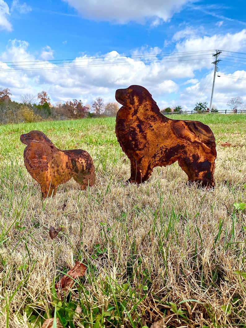 K&ouml;nnte beinhalten: Zwei rostige Metallsilhouetten von Hunden, die auf einer grasbewachsenen Wiese stehen. Der gr&ouml;&szlig;ere Hund steht mit dem Kopf nach links gedreht, w&auml;hrend der kleinere Hund mit dem Kopf nach rechts gedreht steht. Der Himmel ist blau mit wei&szlig;en Wolken.