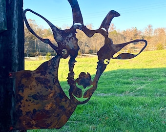 May include: A rusty metal silhouette of a goat's head, mounted on a wooden post. The goat's head is facing to the right, with its ears perked up and its mouth slightly open. The background is a field of green grass.