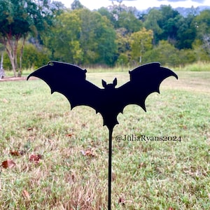 May include: Black metal bat silhouette garden stake with the text "@JuliaRyans2024" on the stake. The stake is in a grassy area with a forest in the background.