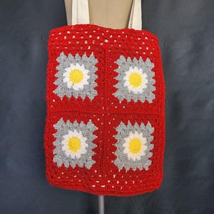 May include: A red crochet tote bag with four daisy-patterned squares. Each square has a grey, white, and yellow flower design. The bag has a cream strap and is displayed on a mannequin. The bag is handmade.