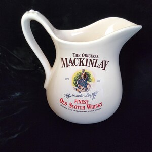 Op de afbeelding: Crèmekleurige keramische kan met een gebogen handvat en tuit. De voorkant heeft de tekst "THE ORIGINAL MACKINLAY FINEST OLD SCOTCH WHISKY" met een illustratie van een man in Schotse kledij. Waarschijnlijk een vintage item.