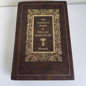 Puede incluir: Un libro de tapa dura marrón con el título "The Complete Works of William Shakespeare" en letras doradas. La portada tiene adornos dorados y la palabra "Illustrated". El libro parece estar en buen estado.