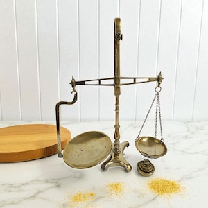 May include: Antique brass balance scale with two oval pans suspended by chains. The scale has a central support and a curved arm. A set of weights and a wooden cutting board are also present.