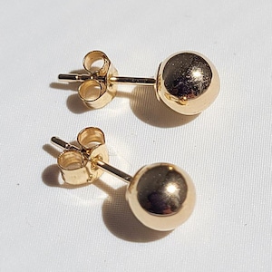 May include: Two gold-colored ball stud earrings. Each earring features a small, round, polished sphere attached to a post with a butterfly backing. The earrings are displayed on a white surface, reflecting light.