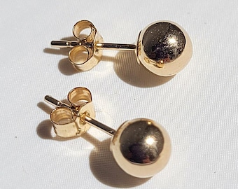Op de afbeelding: Twee goudkleurige bolvormige oorknopjes. Elke oorbel heeft een kleine, ronde, gepolijste bol die is bevestigd aan een pin met een vlindersluiting. De oorbellen worden getoond op een wit oppervlak.