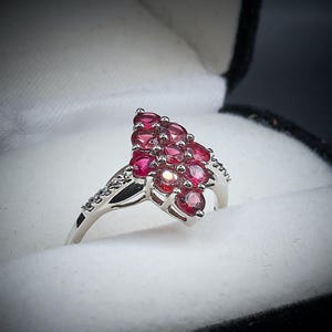 May include: A silver ring featuring a cluster of oval-cut, deep red gemstones. The ring's band is adorned with small, clear accent stones, adding sparkle. The ring is presented in a white jewelry box.