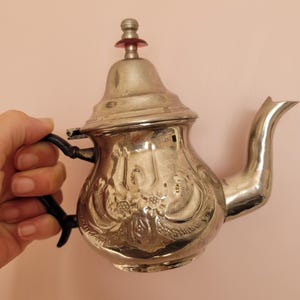 Könnte beinhalten: Eine silberne Teekanne mit einem verzierten Design und einem roten Knopf auf dem Deckel.
