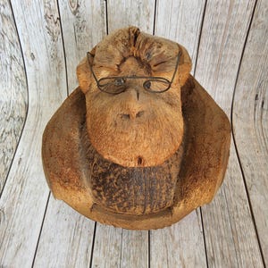 May include: A carved coconut sculpture of a primate wearing wire-rimmed glasses. The sculpture is brown with a textured surface, and the glasses are a thin, silver color. The background is a light-colored wooden surface.