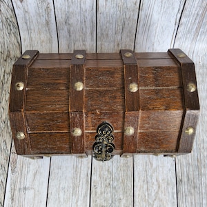 Puede incluir: Un pequeño cofre del tesoro de madera con tinte marrón oscuro. El cofre presenta correas marrones oscuras aseguradas con tachuelas de color latón. Un cierre de metal ornamental asegura la parte delantera. El cofre está sobre una superficie de madera clara.
