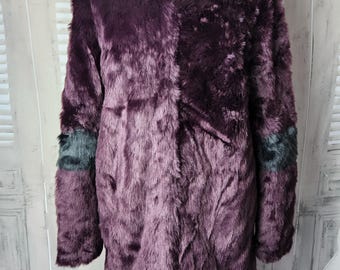 May include: A long, faux fur coat in shades of deep purple. The coat features long sleeves with a dark gray accent on the lower sleeves. The coat is displayed on a mannequin. The coat is a stylish outerwear garment.