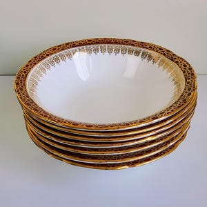 May include: A stack of six white ceramic bowls with a decorative gold and burgundy rim. The rim features a repeating pattern of gold filigree and a burgundy background. The bowls are stacked on a white surface.