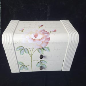 May include: White wooden jewellery box with two drawers and a floral design featuring a pink rose with green leaves. The box has a handwritten inscription on the top and black knobs on the drawers.