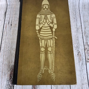 Puede incluir: Un libro antiguo con una ilustración dorada de un caballero con armadura en la portada. El caballero está representado con las manos juntas en oración. El libro tiene un lomo oscuro y un aspecto desgastado.