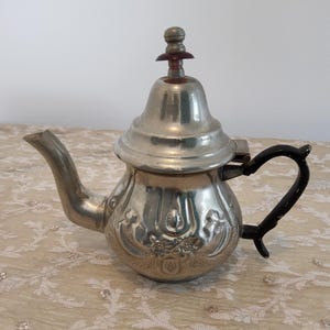 May include: A silver teapot with an ornate design and a black handle. The teapot has a lid with a small knob on top.