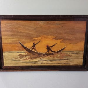 May include: Wooden marquetry artwork depicting two figures in a boat on the water. The artwork features a dark brown frame and a warm color palette of oranges, browns, and tans. The figures are silhouetted, and the waves are detailed.