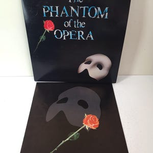 May include: A black vinyl record jacket and inner sleeve for The Phantom of the Opera. The cover features the title in white and blue text, a red rose, and a white mask. The inner sleeve displays a dark mask silhouette and a single red rose.