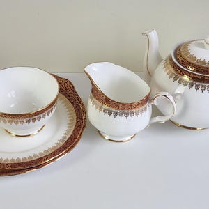 Peut inclure: Service à thé blanc avec des bordures dorées et marron. L'ensemble comprend une théière, un pot à crème, une tasse et une soucoupe. La soucoupe a un bord festonné décoratif. La théière a un couvercle et un bec courbé.