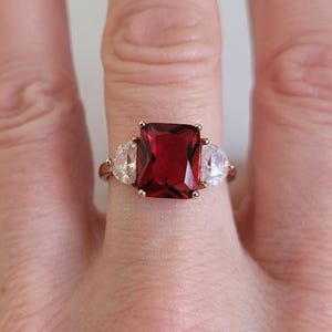 May include: A rose gold ring featuring a large, rectangular, deep red gemstone flanked by two smaller, pear-shaped, clear gemstones. The ring's band is a delicate rose gold color, and the gemstones are set in a classic, elegant design.