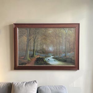 Puede incluir: Pintura enmarcada de un paisaje forestal con un arroyo. La obra presenta un sendero, árboles y un río que fluye. La pintura está en un marco de madera marrón oscuro y cuelga de una pared beige.