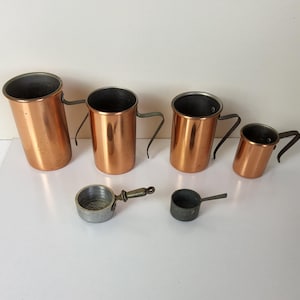 Pode incluir: Um conjunto de xícaras medidoras e colheres de cobre. As xícaras, de tamanhos variados, possuem alças curvas e um acabamento brilhante. Duas pequenas colheres de metal também estão presentes, uma com cabo.
