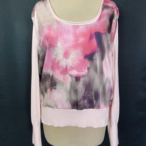 May include: A light pink long-sleeved sweater with a scoop neck. The front features a sheer overlay with a large floral design in shades of pink, purple, and white. The sweater has a ribbed hem and cuffs.