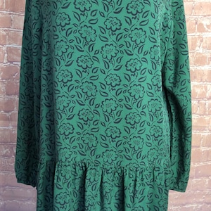 May include: A long-sleeved dress in a vibrant green hue featuring a repeating dark charcoal floral pattern. The garment has a round neckline and a gathered drop-waist seam that creates a subtle ruffle effect along the lower portion of the skirt.