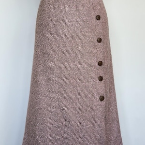 May include: A long, mauve tweed skirt with a button-up closure. The skirt features a high waist and a slight A-line shape. The buttons are a dark brown colour. The fabric has a textured appearance.
