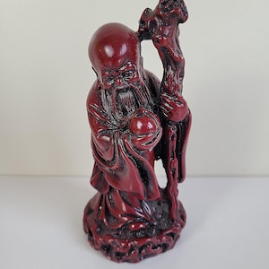 Può includere: Statuetta rossa di un uomo anziano con una lunga barba, che tiene un bastone e un oggetto rotondo. La statuetta presenta dettagli intricati ed è ambientata su uno sfondo semplice. La figura indossa una veste.