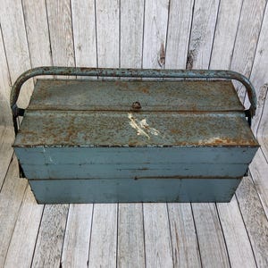 May include: A vintage blue metal toolbox with a curved handle. The toolbox has a rectangular shape with a hinged lid and multiple storage compartments. The metal shows signs of rust and wear, giving it a rustic appearance. Designed for storing tools.