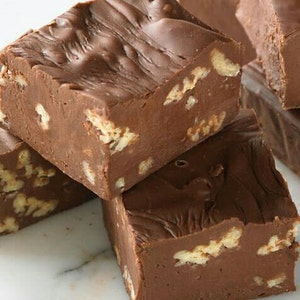 May include: Close-up of a stack of chocolate fudge squares with pecans. The fudge is a rich brown color and has a smooth, glossy surface. The pecans are scattered throughout the fudge and add a crunchy texture.