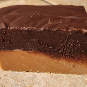 May include: A square piece of fudge with a layer of dark chocolate on top. The fudge is a light brown color and the chocolate is a dark brown color.