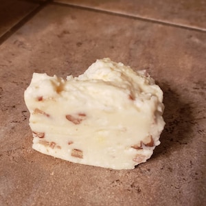 May include: A close-up of a piece of white fudge with visible pecan pieces. The fudge has a creamy texture and a slightly uneven surface. The pecans are scattered throughout the fudge.