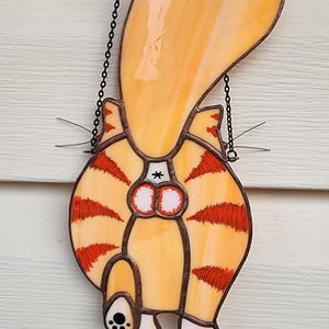 May include: Stained glass cat butt hanging decoration. The orange and yellow cat has red stripes and a black chain for hanging. The cat's tail is arched upwards.