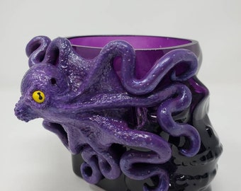 Peut inclure: Crâne en verre violet avec une pieuvre violette enroulée autour. La pieuvre a un œil jaune.