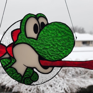 May include: A stained glass window hanging ornament featuring a green and red cartoon character with a long red tongue. The character has a large green eye and a textured green shell.