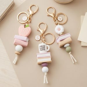 May include: Three gold-toned metal keychains featuring silicone beads, miniature book stacks, and charms. One keychain has a pink apple charm with the word TEACH, one has a white coffee mug with the text coffee teach repeat, and one has a white daisy flower.