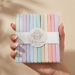 Puede incluir: Una mano sostiene un paquete de artículos de papelería rectangulares de colores pastel. Los artículos están dispuestos en filas de varios colores, incluyendo azul claro, verde, rosa y lavanda. El paquete está envuelto con una banda blanca que tiene el texto "The New Season Shop".