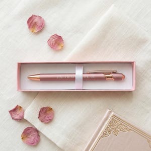 May include: A rose gold pen with the words "FAITH OVER FEAR" in a pink gift box. The pen is displayed on a white cloth with scattered dried rose petals and a pink journal.