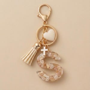 May include: A gold-toned keychain featuring a letter S charm filled with small beige and white stone-like fragments. The keychain includes a cream-colored tassel, a small white heart charm, and a small white cross charm attached to a gold ring.