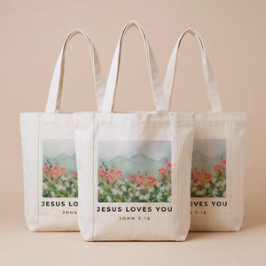 May include: Three off-white canvas tote bags featuring a rectangular graphic print of a watercolor-style landscape with pink and white flowers against green hills. The text "JESUS LOVES YOU" and "JOHN 3:16" is printed in black below the illustration.