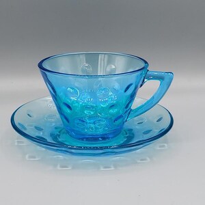 May include: A clear, light blue teacup and saucer set. The cup and saucer have a textured, bubble-like pattern. The cup has a handle and sits on a matching saucer. The set is made of glass and is suitable for serving tea or coffee.