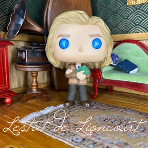 May include: A vinyl figure of Lestat de Lioncourt with blonde hair and bright blue eyes, wearing a tan cardigan over a white shirt and dark trousers. The figure holds a small green book with both arms crossed in front of the chest.