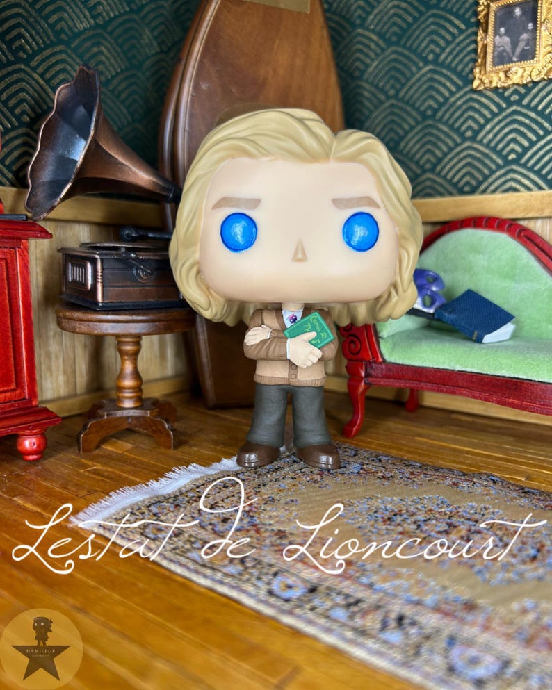 May include: A vinyl figure of Lestat de Lioncourt with blonde hair and bright blue eyes, wearing a tan cardigan over a white shirt and dark trousers. The figure holds a small green book with both arms crossed in front of the chest.