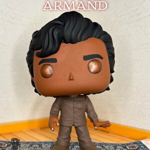 May include: A vinyl collectible figure of The Vampire ARMAND with dark wavy hair, brown skin, and metallic copper eyes. The figure wears a brown button-down shirt and matching trousers. The text The Vampire ARMAND is displayed above the figure.